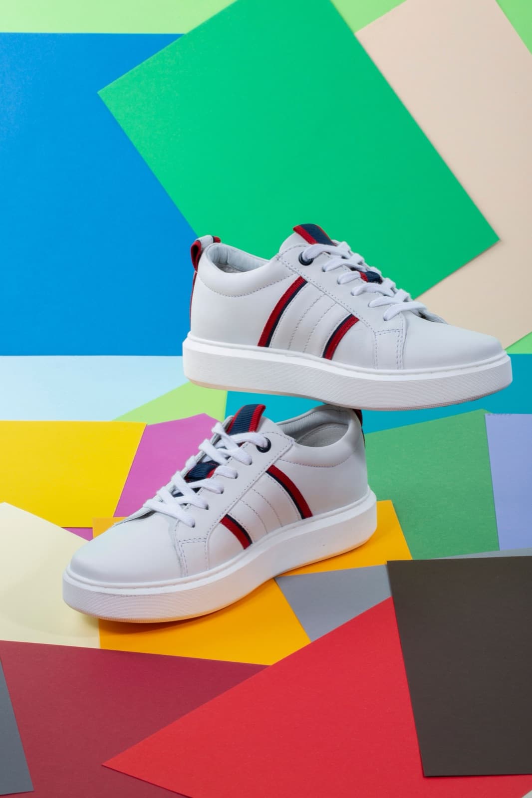 A pair of white striped sneakers side by side on green, blue and red paper