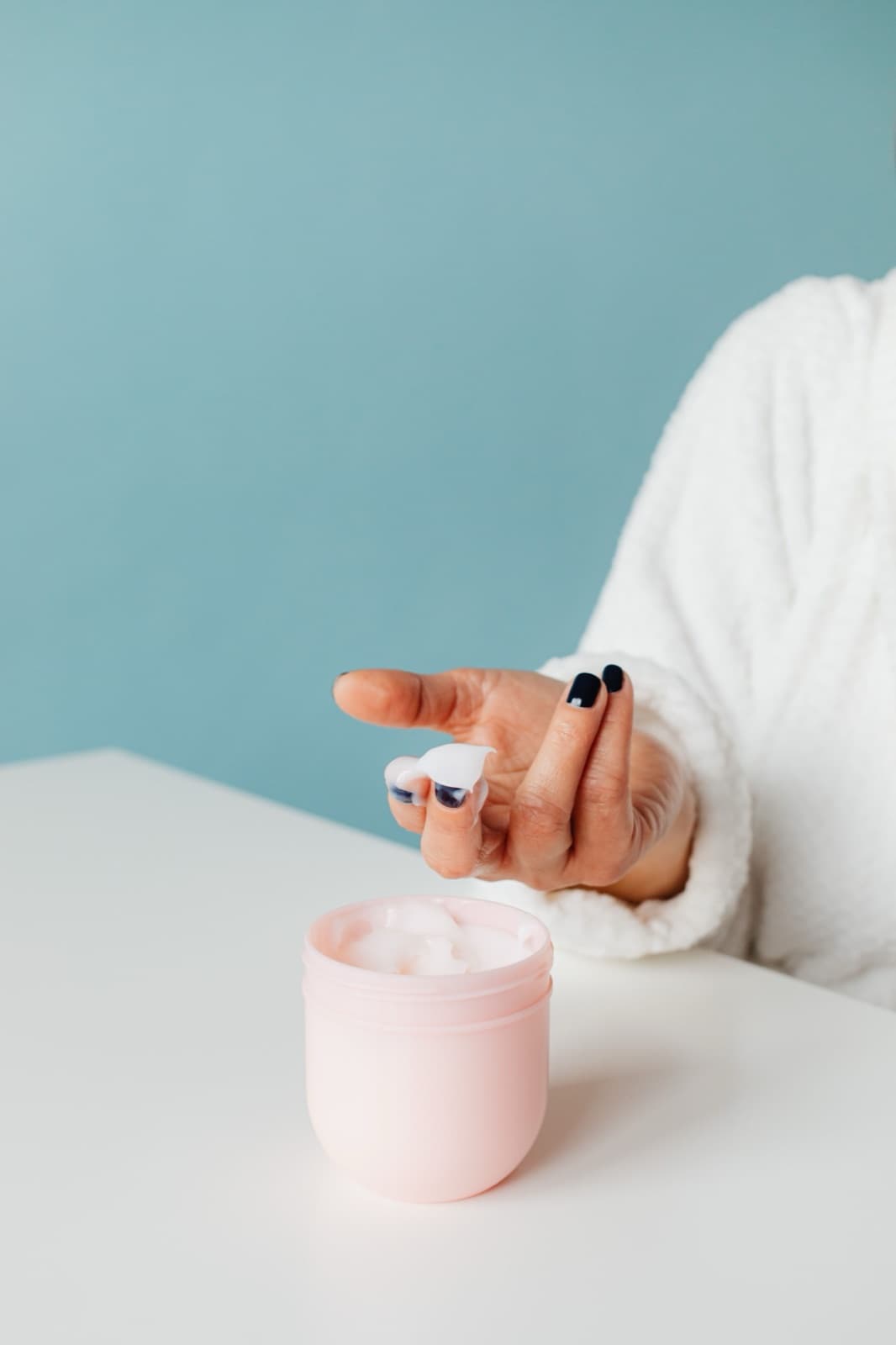 An open pink cream jar on a white table, a hand holding a dab of cream