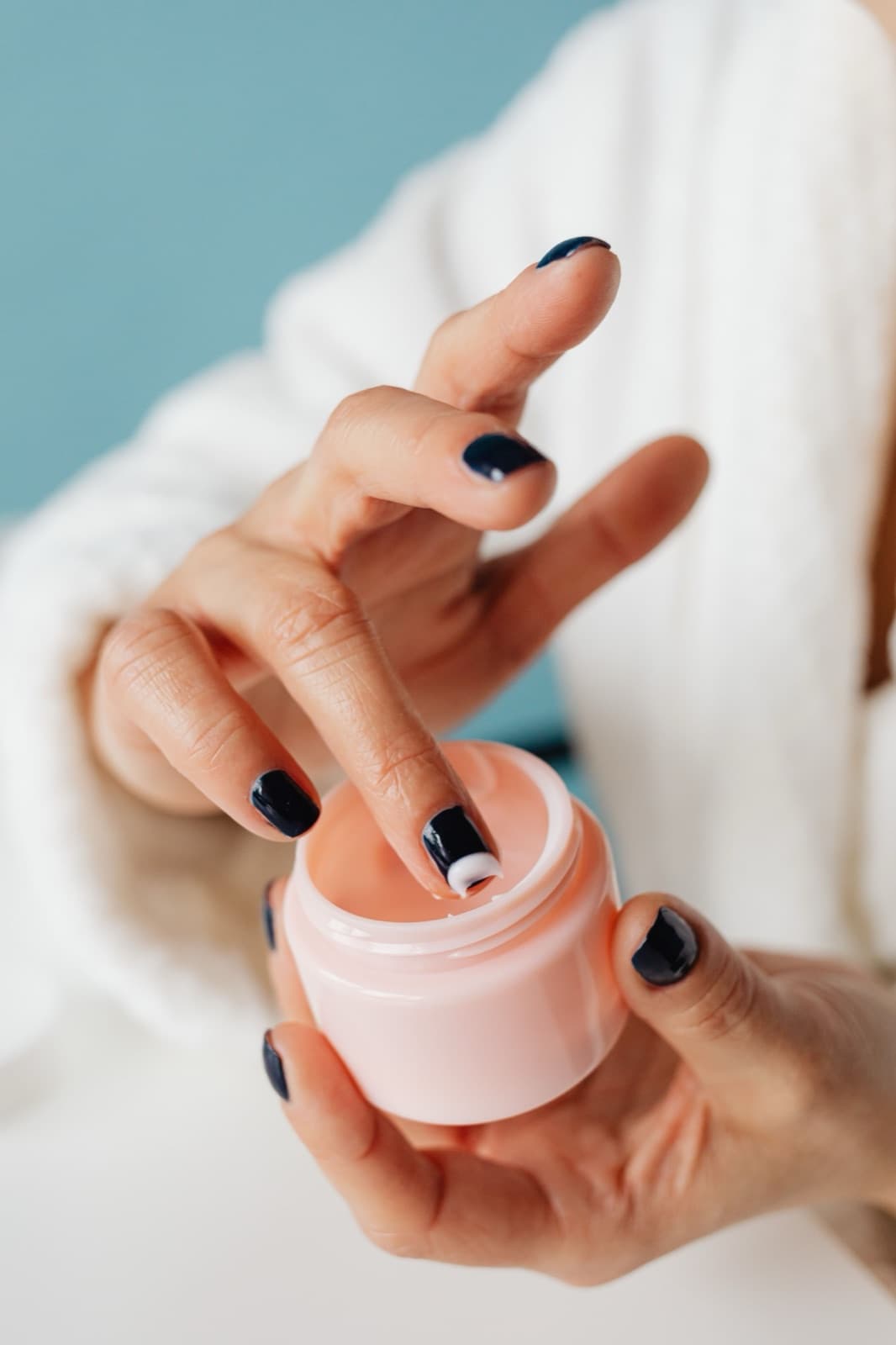 A finger dipping into the pink jar of whipped cream against a light blue wall