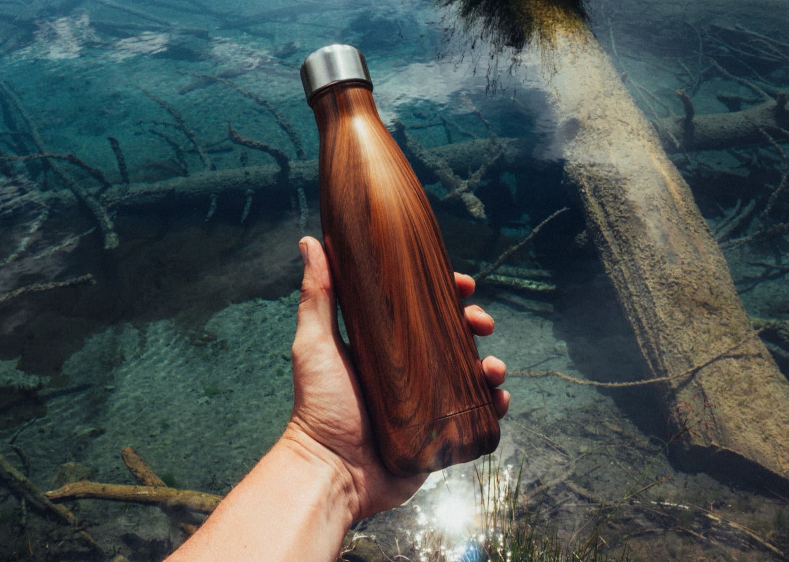 A hand holding the wood-grain bottle over a clear mountain stream